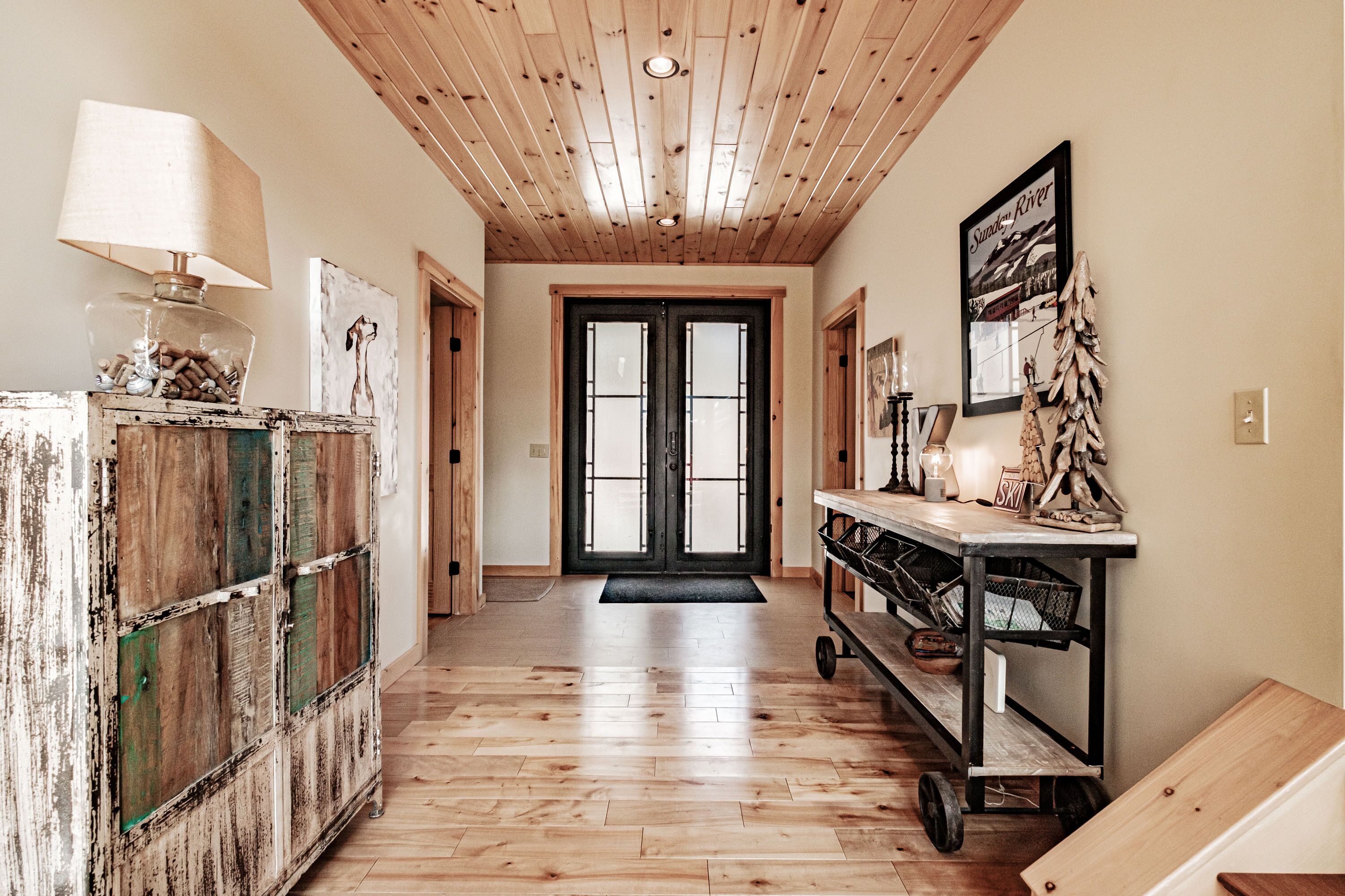 A hallway with a wooden floor, a rustic cabinet, a lamp, a framed picture, and a cart with baskets.