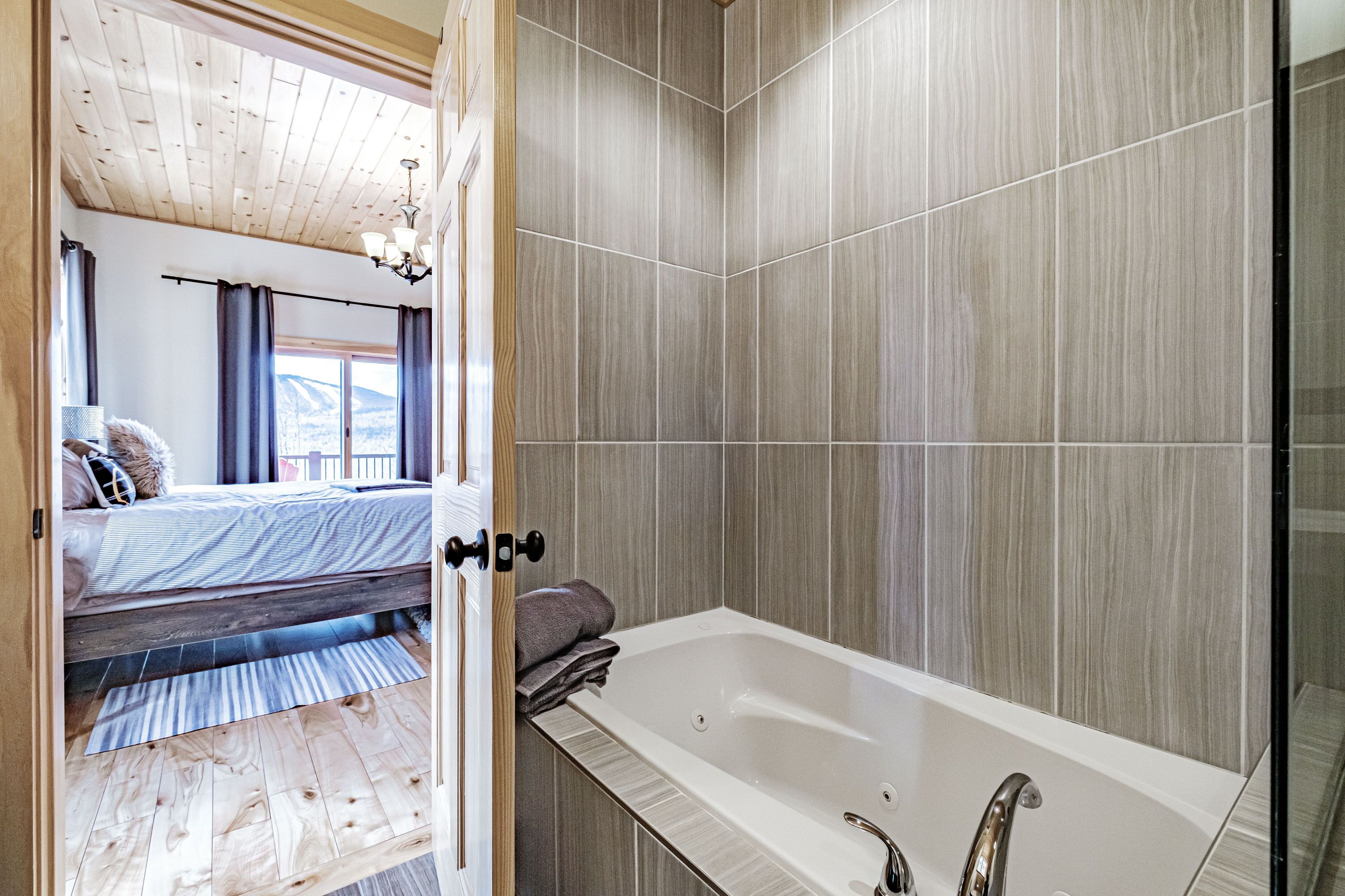 A bedroom with a wooden ceiling, a bed, a window with curtains, and a bathroom with a bathtub and tiled walls.