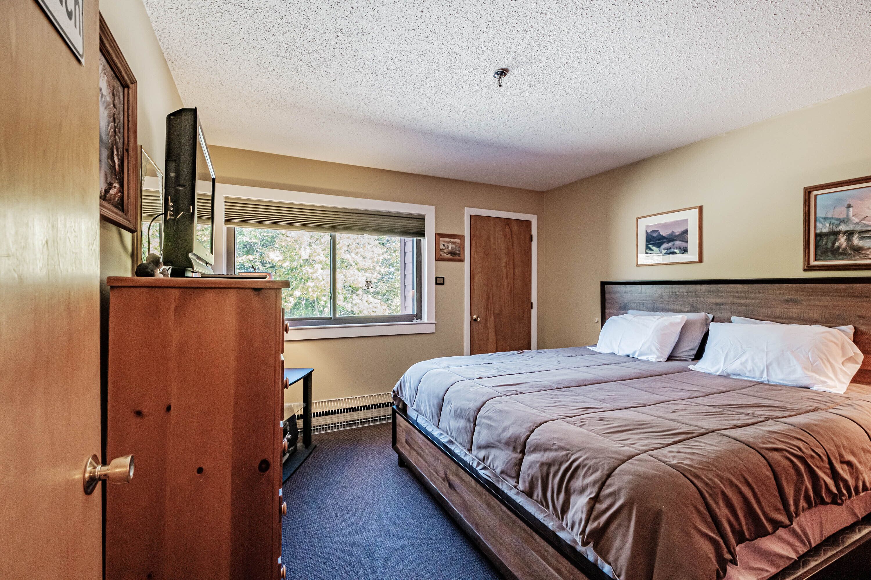 A bedroom with a large bed, a wooden dresser, a desk, and a window with blinds.