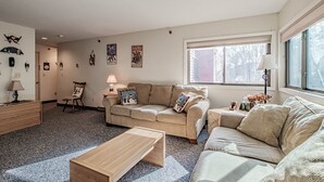 A living room with a beige sofa, a wooden coffee table, and a lamp. There are wall decorations and a window with blinds. - On Mountain two bedroom unit sleeping 6 (Newry)