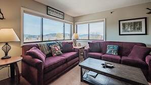 A living room with two purple sofas, a wooden coffee table, and a view of the outdoors through large windows. - On Mountain two bedroom unit sleeping 8 (Newry)
