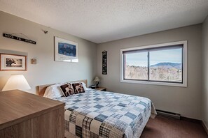 A bedroom with a large window, a bed with a checkered quilt, a nightstand, a lamp, and wall decorations including a framed picture and a sign.