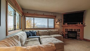 A living room with a sectional sofa, a fireplace, and a large window with a view of trees and mountains. - On Mountain two bedroom unit sleeping 6 with air conditioning! (Newry)
