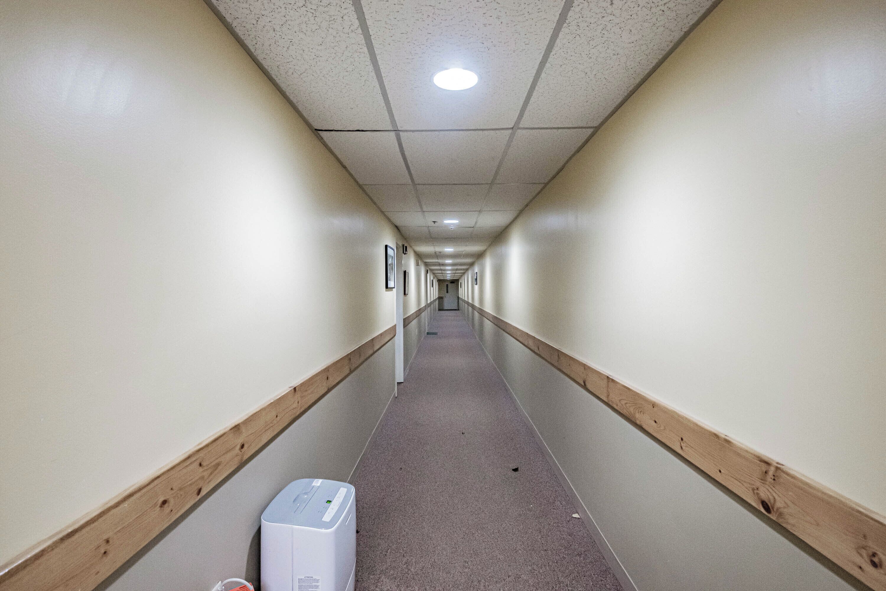 A long, narrow hallway with wooden baseboards, a white wall, and a light-colored floor.