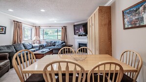 A cozy living room with a wooden dining table, comfortable seating, a fireplace, and a large window with curtains. - North Peak 2-305 Ski-in Ski-out (Newry)