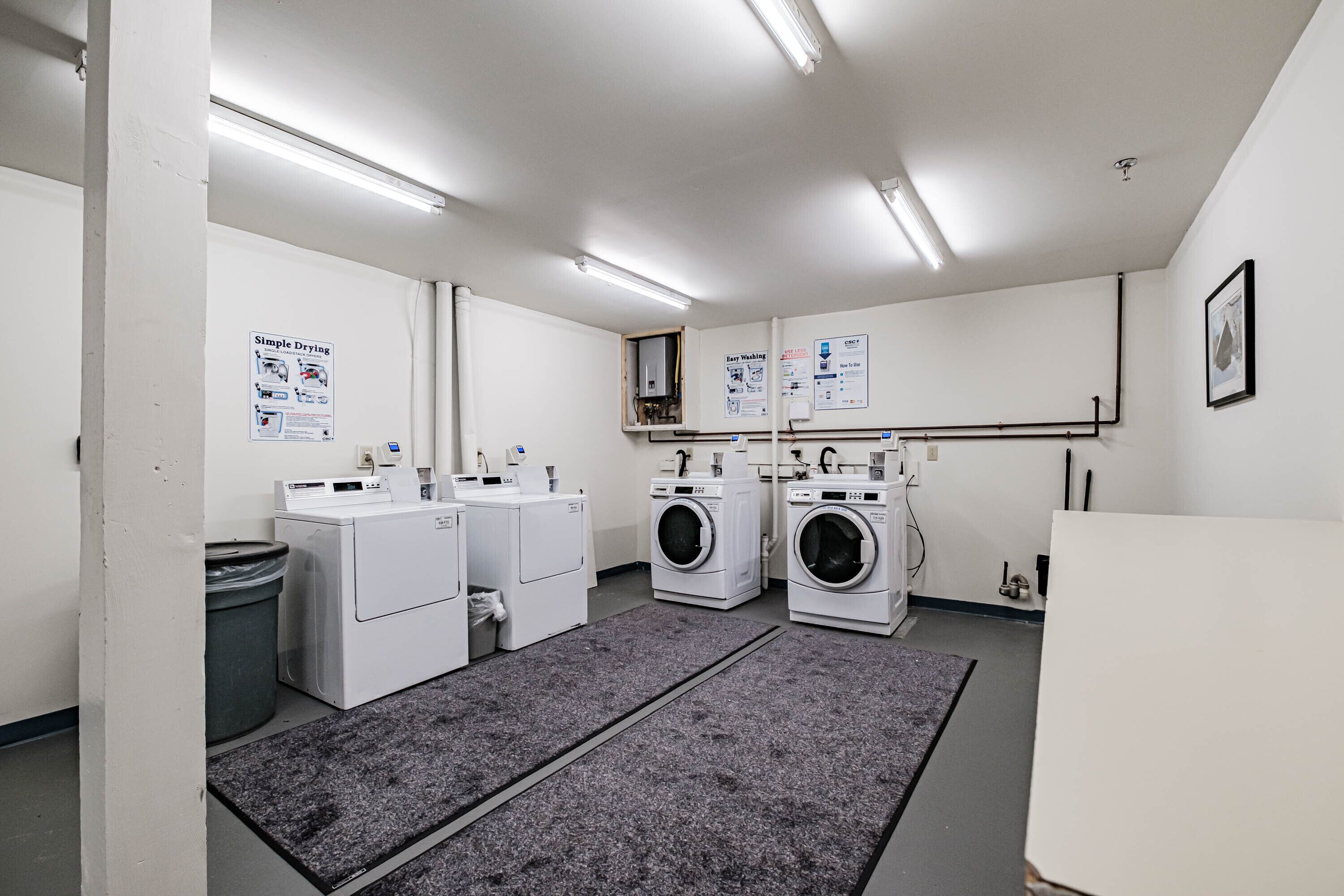 A laundry facility with four washing machines, a trash bin, and informational posters on the walls.