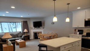 Une cuisine moderne avec des meubles blancs, un îlot central doté de tabourets, un coin repas avec des chaises en bois, une cheminée et un téléviseur fixé au mur.