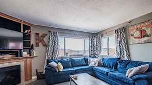 A living room with a sectional sofa, a flat-screen TV, a fireplace, and a large window with curtains.