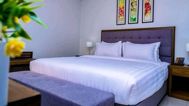 A neatly arranged bedroom with a large bed, a bedside table, a nightstand, and a bench. - Beautiful woman hotel (Kampala)