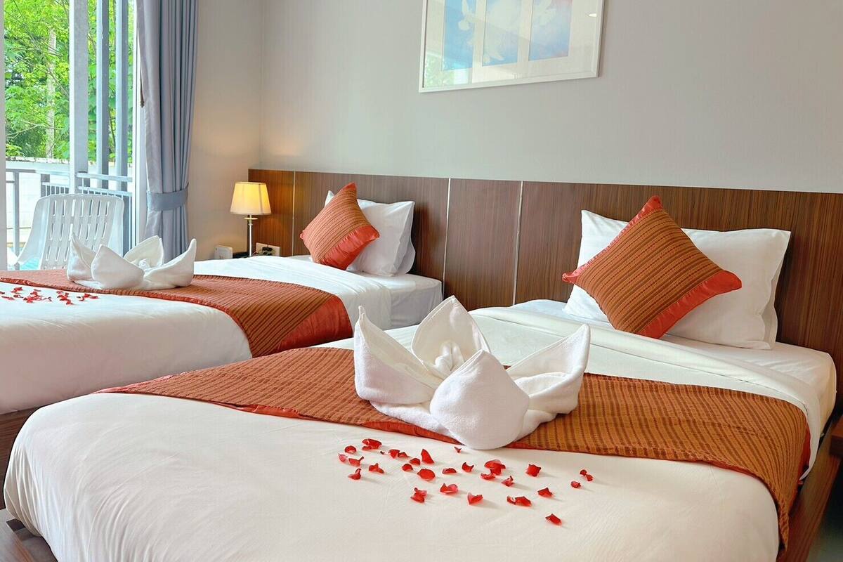 Ein Hotelzimmer mit zwei Betten, jeweils mit weißer Bettwäsche und roten Akzenten, einem Holzkopfstück, einem Fenster mit Vorhängen und einem gerahmten Bild an der Wand.