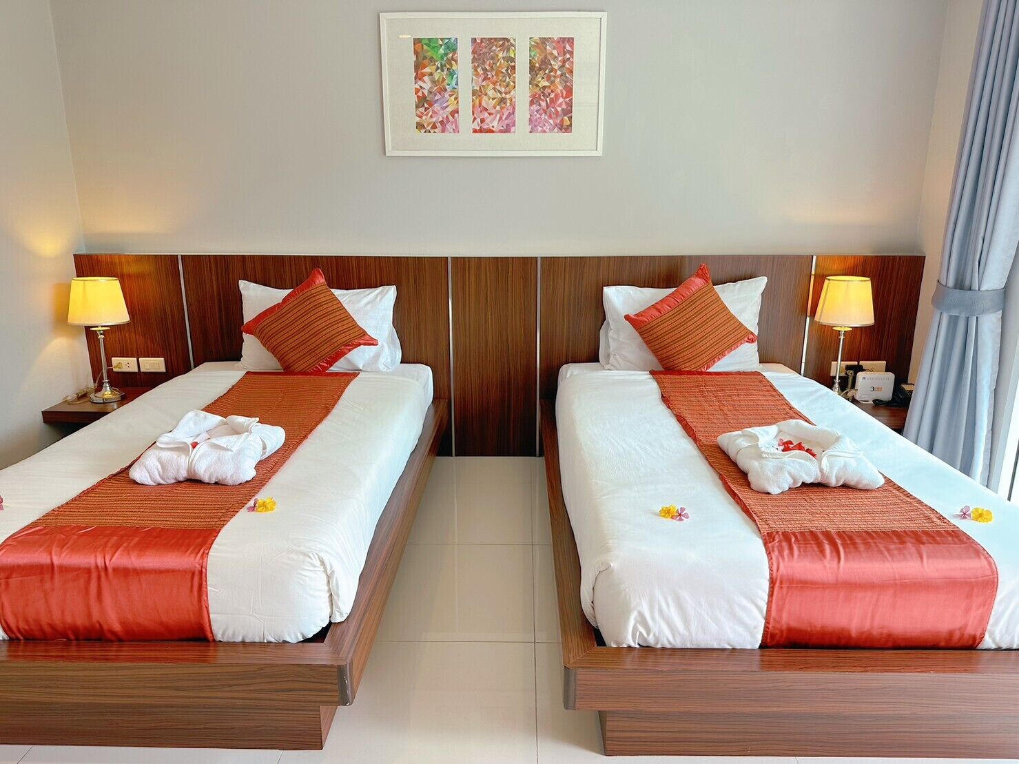 Habitació d'hotel amb dos llits, cadascun amb fusta al capçal, roba de llit blanca i detalls vermells. Hi ha dues làmpades de nit, una obra d'art emmarcada sobre els llits i una finestra amb cortines.
