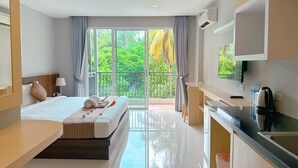 Une chambre d’hôtel moderne équipée d’un lit, d’un bureau, d’une kitchenette et d’un balcon offrant une vue sur la verdure.