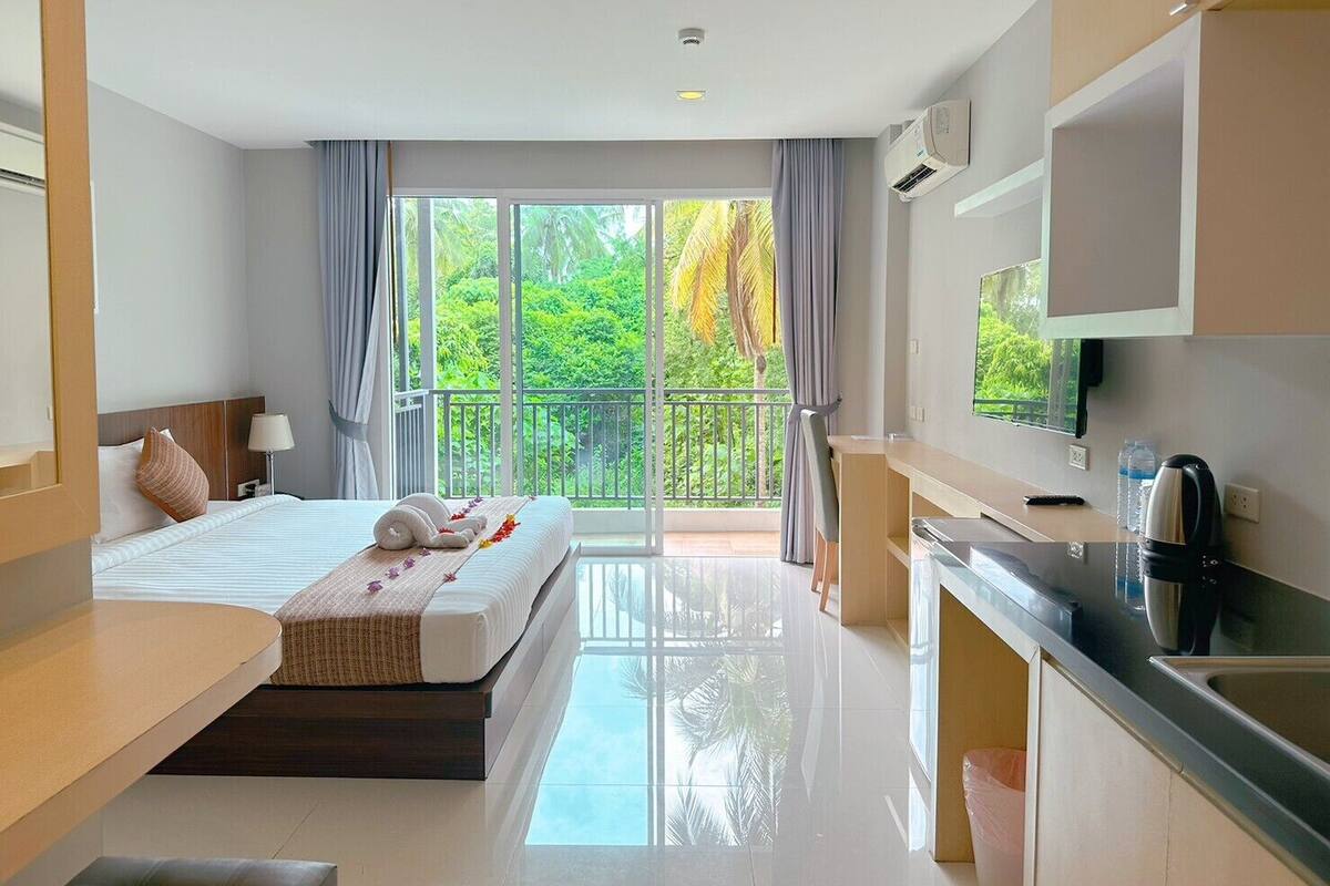 Ein modernes Hotelzimmer mit Bett, Schreibtisch, einer Küchenzeile und einem Balkon mit Blick auf Grünflächen.