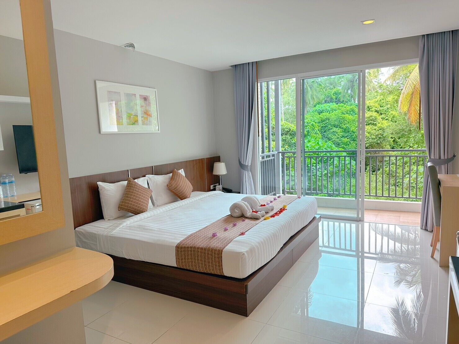 Une chambre d’hôtel avec un grand lit, une table de chevet, un miroir et une vue sur une végétation luxuriante depuis le balcon.