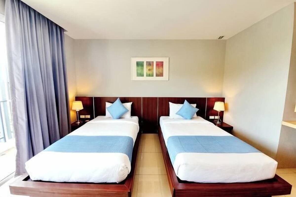 Ein Hotelzimmer mit zwei Betten, einem Nachttisch mit Lampe, einem Fenster mit Vorhängen und einem an der Wand hängenden Bild.
