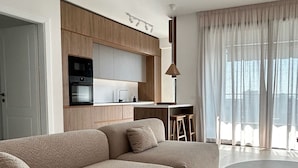 A modern living room with a large beige sectional sofa, a round coffee table, and a kitchen area with wooden cabinets and a dining table.