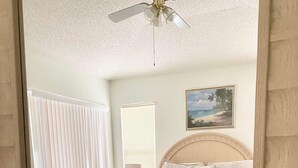 Un dormitorio con cama, mesita de noche, lámpara, ventilador de techo y ventana con persianas.