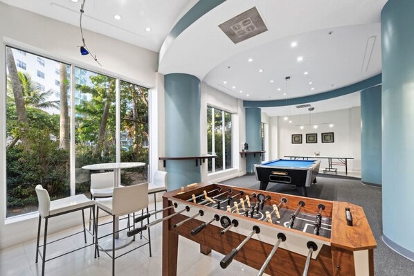 Un espace de loisirs intérieur moderne, doté d’un baby-foot, d’une table de billard et d’un coin salon avec de grandes fenêtres donnant sur un paysage verdoyant.