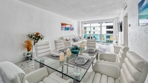 A modern dining area with a glass table, white furniture, and a view of buildings through large windows. - Beachfront Condo| Steps to Broadwalk | Sleeps 6|Pool+Gym (Hollywood)