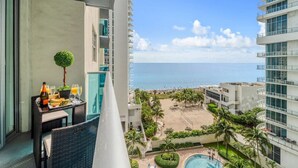 A balcony with a table set for breakfast, a small tree, and a view of the ocean and beach. - Beachfront Condo| Steps to Broadwalk | Sleeps 6|Pool+Gym (Hollywood)
