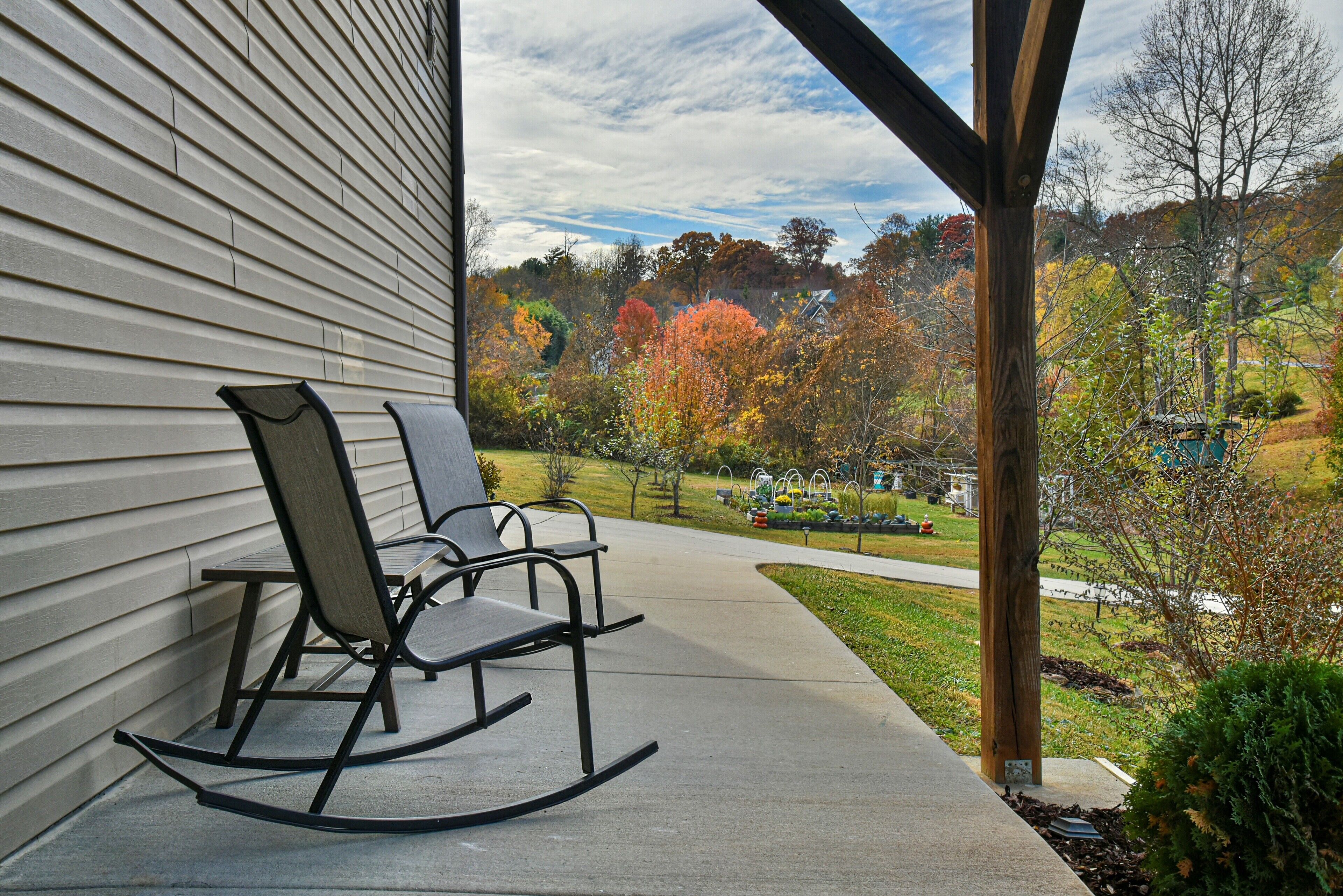 A porch with two rocking chairs, a concrete pathway, and a view of a park with autumn trees.
