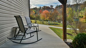 A porch with two rocking chairs, a concrete pathway, and a view of a park with autumn trees.