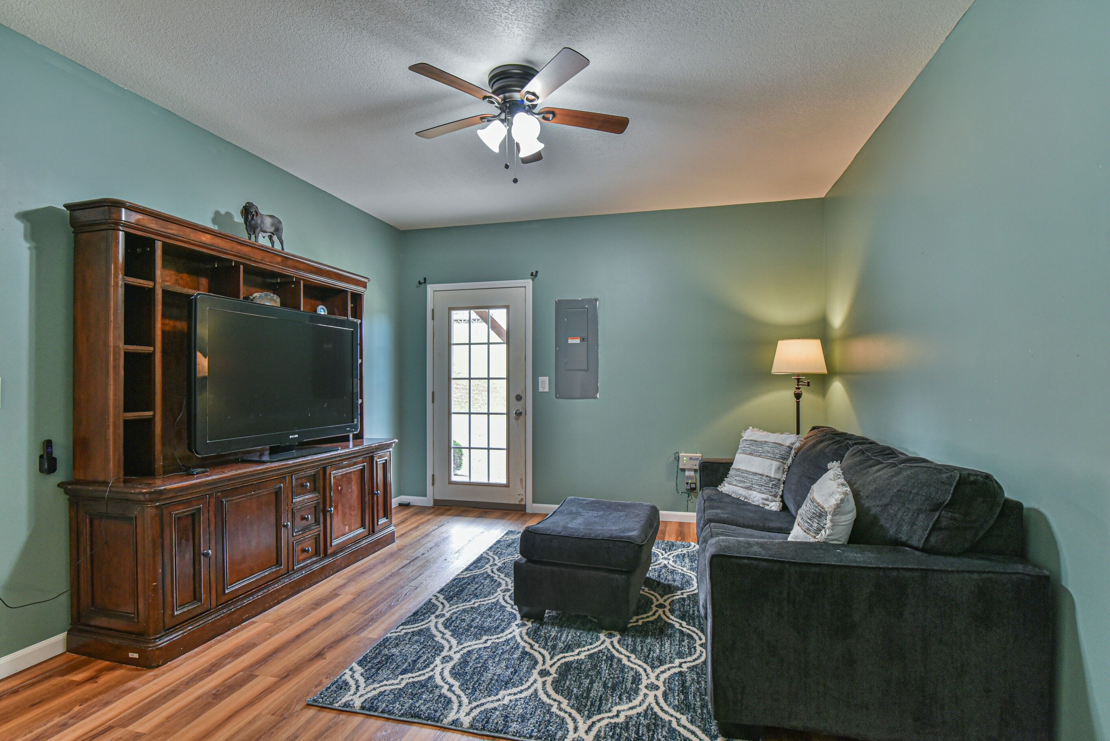A living room with a wooden entertainment center, a flat-screen TV, a ceiling fan, a dark-colored sofa, and a patterned rug.