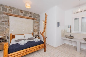 Quarto com parede de pedra, cama de madeira, mesa de cabeceira e espelho, além de uma janela.