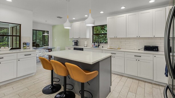 Una cocina moderna con armarios blancos, una isla central con taburetes y luminarias colgantes, y vista a zonas verdes.