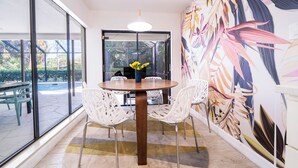 Un comedor con una mesa redonda, sillas blancas, un florero con flores amarillas y un gran mural tropical en la pared.