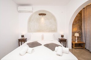 A hotel room with a bed, two bedside tables, an air conditioning unit, and a stone wall. - Peristeronas Village (Mykonos)