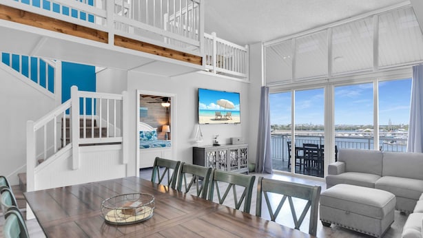 A spacious living area with a wooden dining table, white railings, and a large window offering a view of the water.