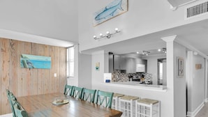 A dining area with a wooden table and chairs, a kitchen in the background, and artwork on the walls.