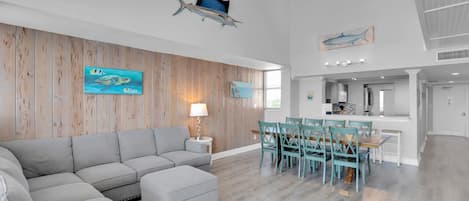 Un vaste espace de vie comprenant un grand canapé d’angle gris, une table à manger avec des chaises de couleur turquoise et un lambris en bois sur les murs.