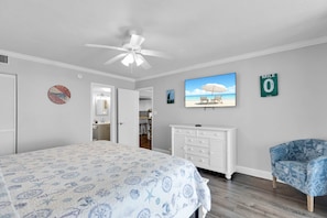 Une chambre à coucher comprenant un lit, un ventilateur de plafond, une commode, une chaise et un mur orné d’un tableau représentant une plage.