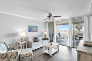 Un salon comprenant un canapé, deux fauteuils, une table basse et un ventilateur de plafond. Un tableau est accroché au mur, et on peut voir, par la fenêtre, un patio extérieur avec une table et des chaises.
