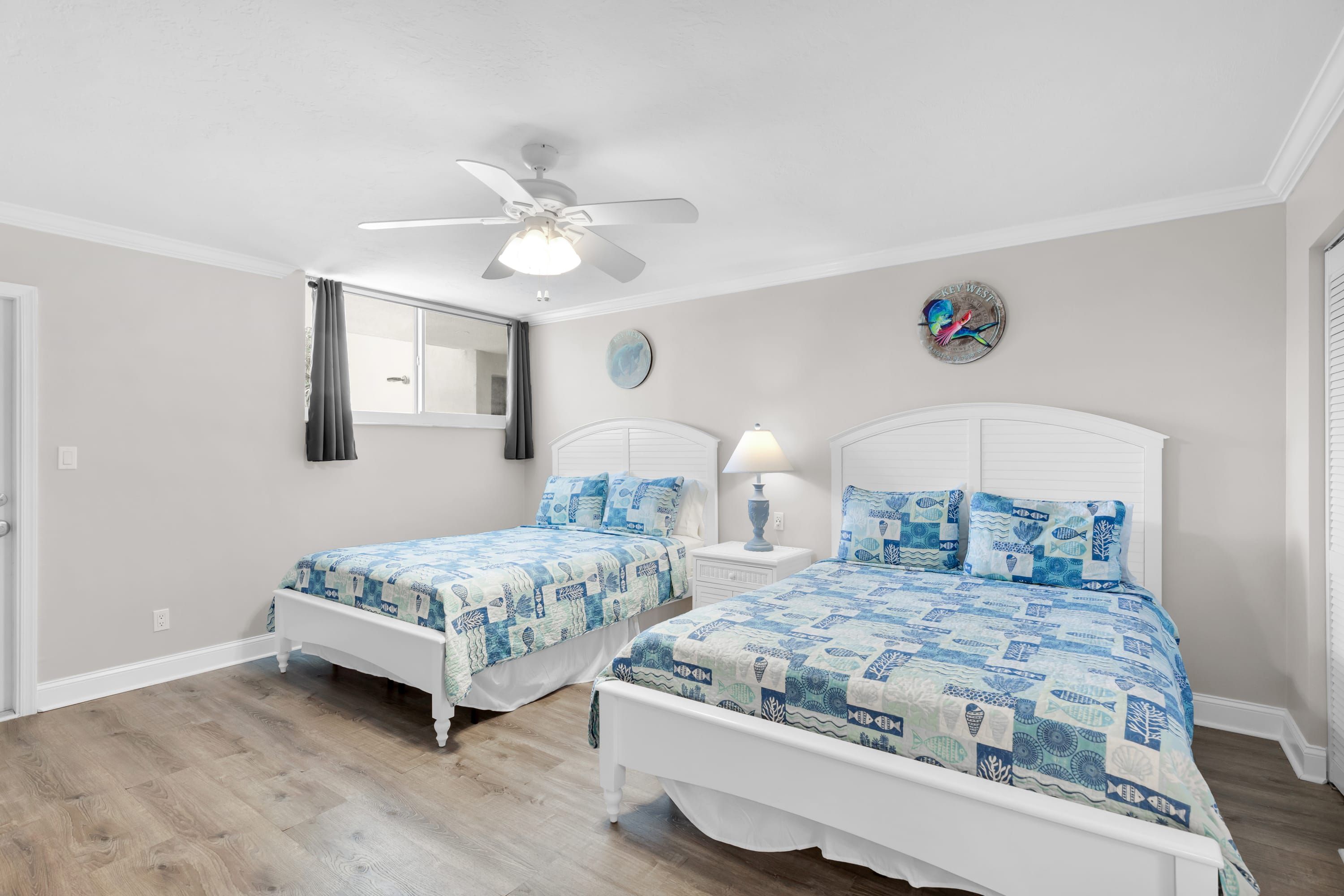 A bedroom with two beds, a ceiling fan, a window with curtains, a wall clock, and a decorative plate.