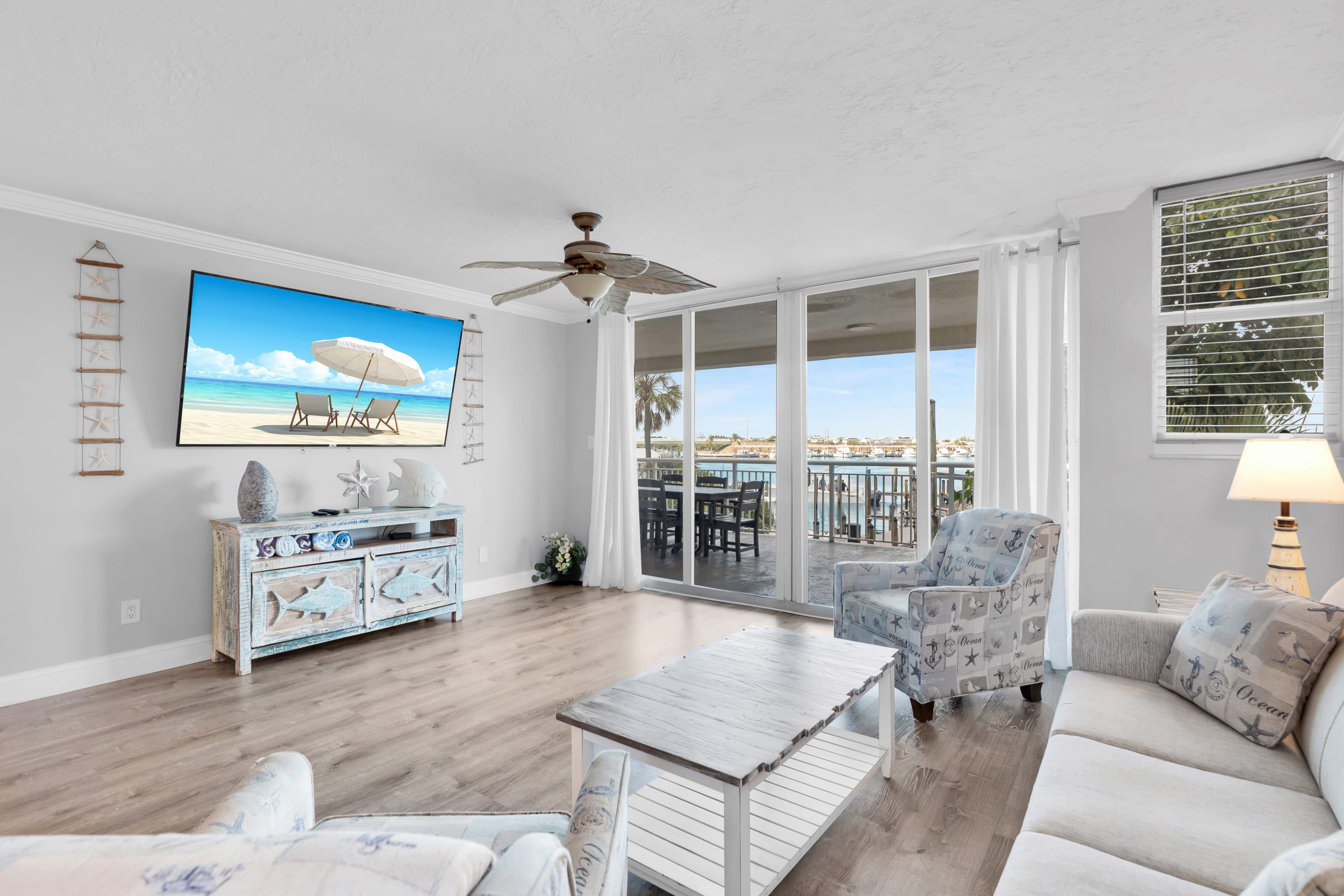 A spacious living room with a large TV, a ceiling fan, and a view of a pool and palm trees through the glass doors.