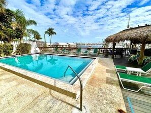 A swimming pool with a clear blue water, surrounded by lounge chairs and a thatched roof structure. - Waterfront Condo | Pool, Dock, Parking, Sleeps 8 (Key West)