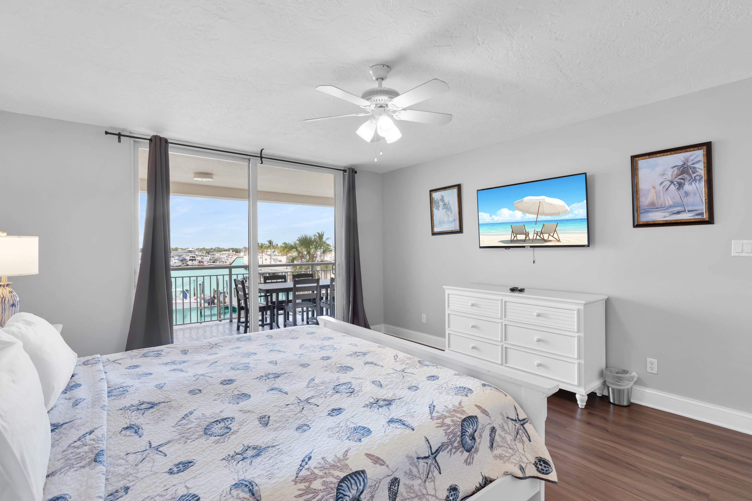 A bedroom with a bed, a dresser, a ceiling fan, a TV, and a balcony with a view of the marina.