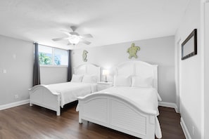A bedroom with two beds, a ceiling fan, a window with curtains, and a wall-mounted mirror.