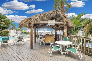 A wooden deck with a thatched roof structure, a pool, lounge chairs, and a bar area. - Waterfront Condo | Pool, Dock, Parking, Sleeps 8 (Key West)
