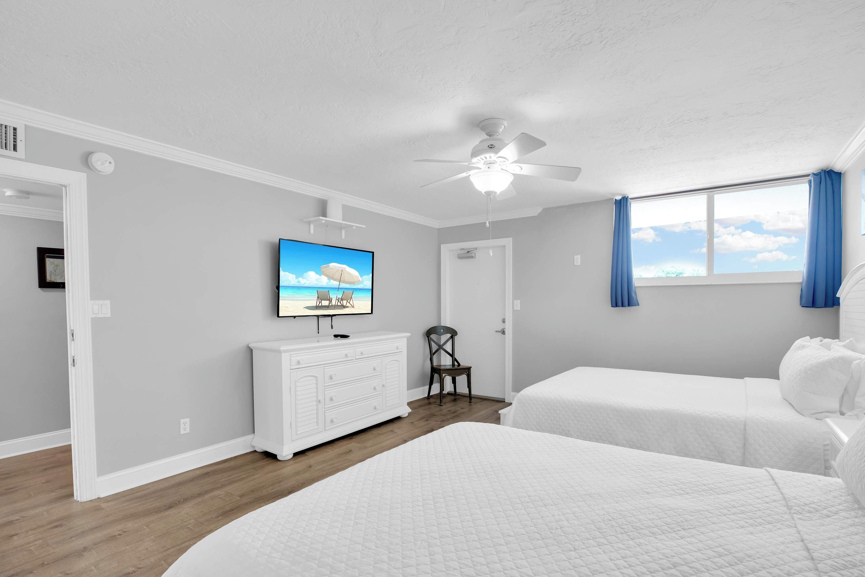 Ein Schlafzimmer mit zwei Betten, einem Deckenventilator, einem an der Wand befestigten Fernseher und einem Fenster mit Vorhängen.