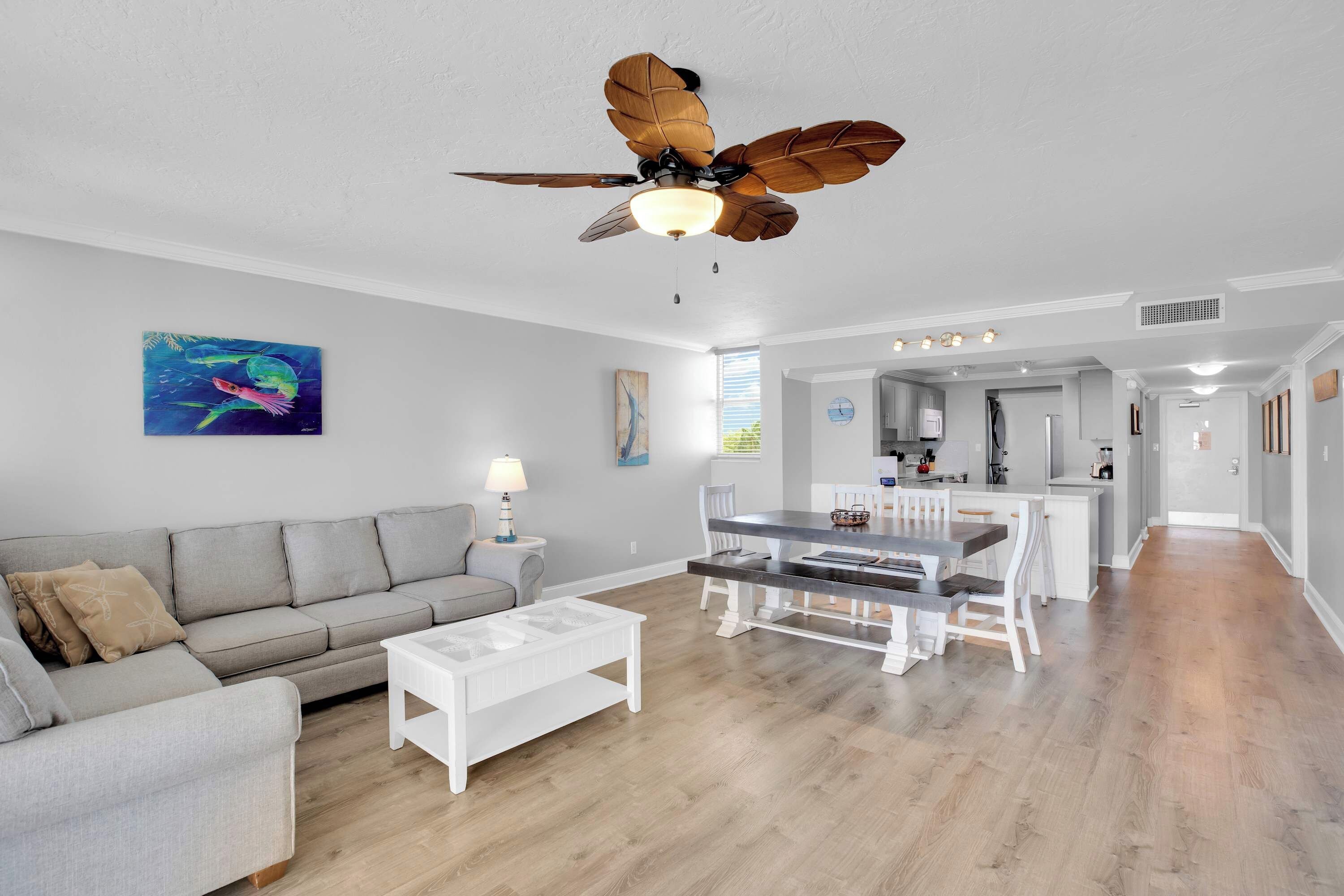 A modern living room with a gray sofa, a white coffee table, and a dining area with a wooden table and chairs. A ceiling fan with wooden blades is visible.
