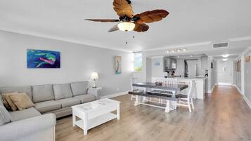 A modern living room with a gray sofa, a white coffee table, and a dining area with a wooden table and chairs. A ceiling fan with wooden blades is visible.