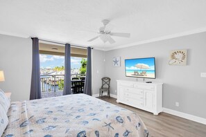 A bedroom with a bed, a TV, a ceiling fan, and a balcony with a view of the beach.