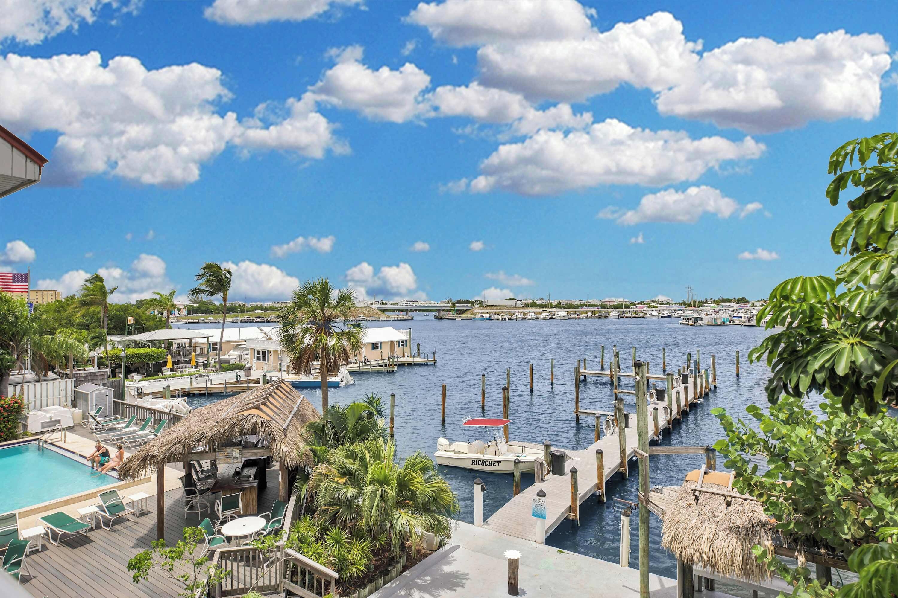 A marina with docked boats, a swimming pool with lounge chairs, and a view of the water and distant buildings.