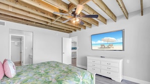 A bedroom with a bed, a ceiling fan, a white dresser, and a beach-themed TV on the wall.