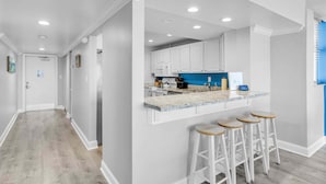 A modern kitchen with white cabinets, a blue backsplash, and a marble countertop. There are four wooden stools at the counter.