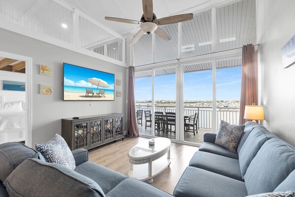 A spacious living room with a large TV, a ceiling fan, and a view of the sea through large windows.